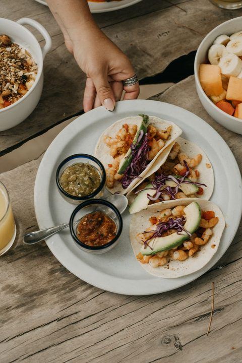 A close-up of hand holding a plate of tacos, garnished with avocado and red cabbage, served with sal