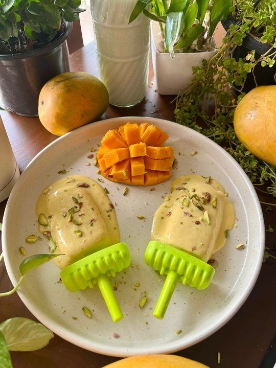 Mango kulfi popsicles served with pistachios and fresh mango slices on a plate.