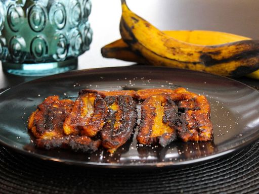 Fried plantains served on a black plate with fresh bananas.